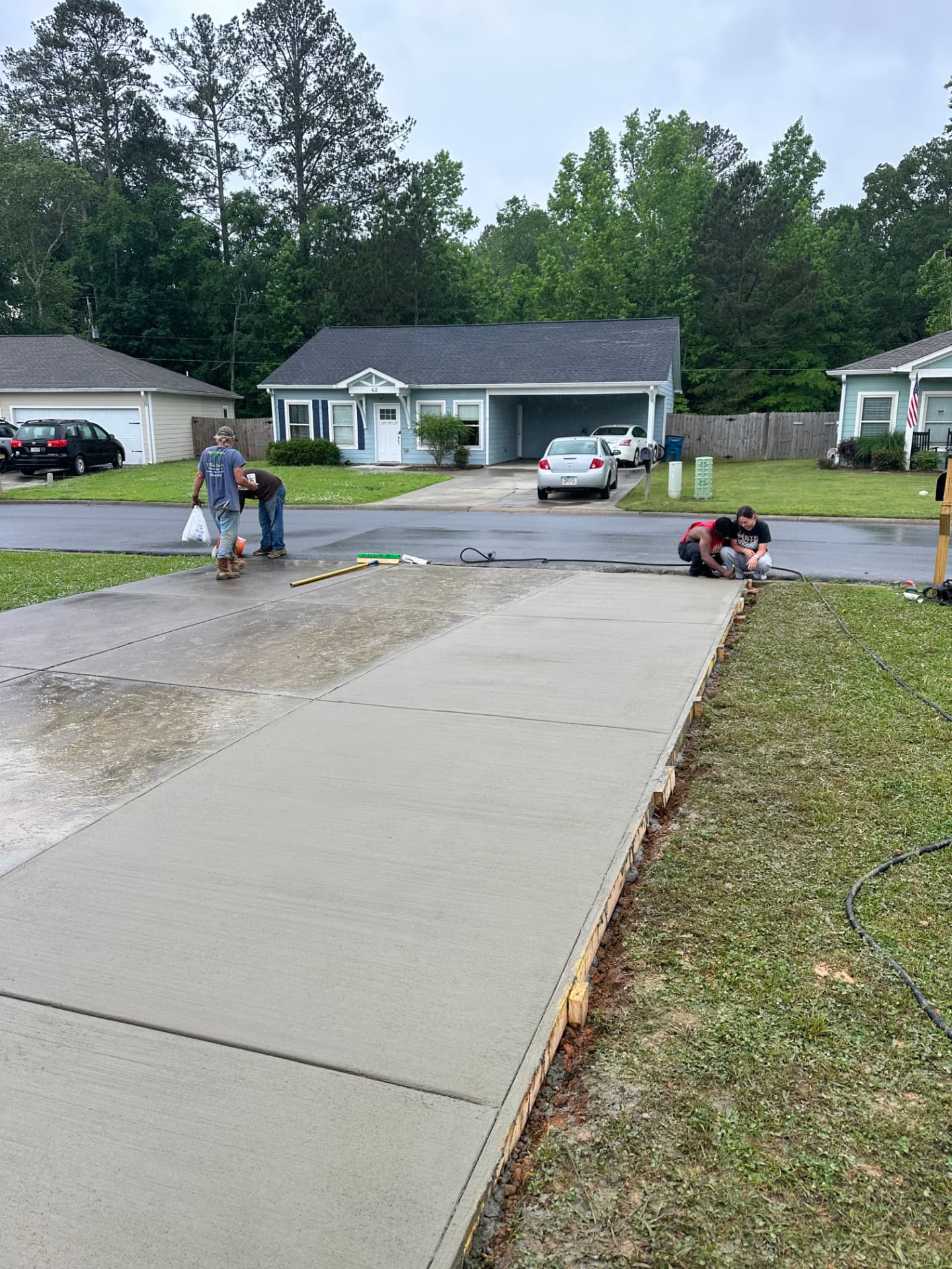 Crew finishing a freshly poured concrete driveway extension in a suburban neighborhood, with wooden forms and a broom-finish surface