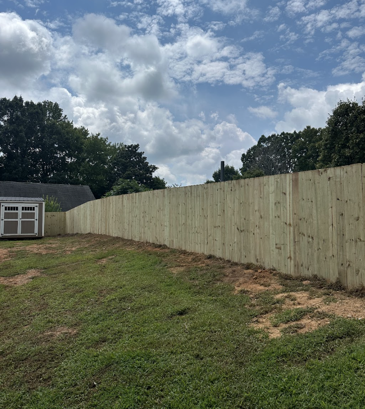 Newly installed wood privacy fence running across a sloped backyard, with a small shed at the corner and trees beyond