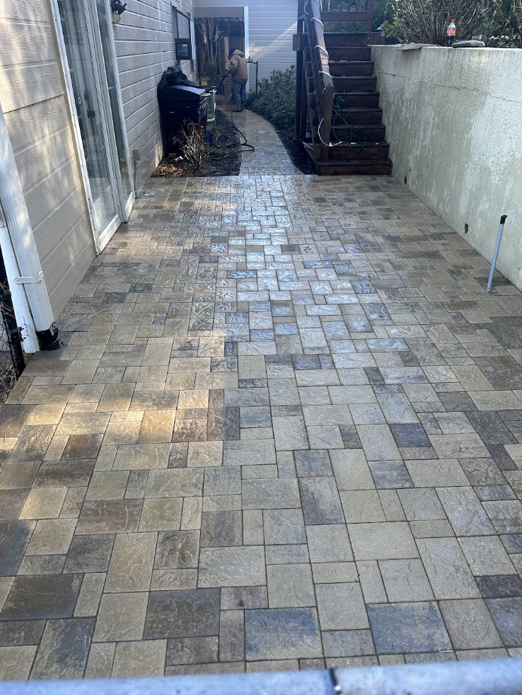 Newly installed stone paver patio in tan, brown, and charcoal tones running between a home and a side wall, being pressure-washed