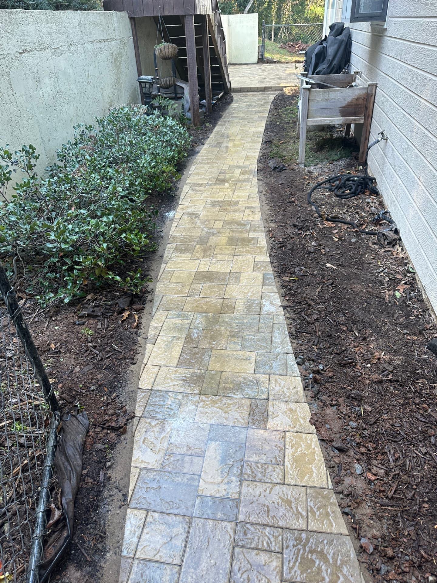 Newly installed paver walkway in tan, beige, and gray tones curving through a narrow residential side yard with mulched beds and a stucco wall